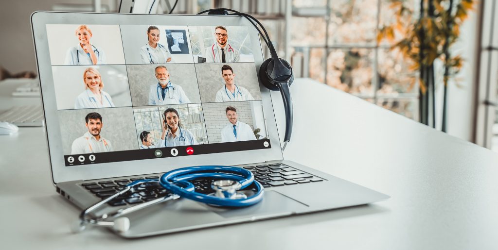 Image of open laptop showing a multi-video conference call in progress, headphones draped over the side of the screen, and a stethoscope sitting on top of the keyboard.